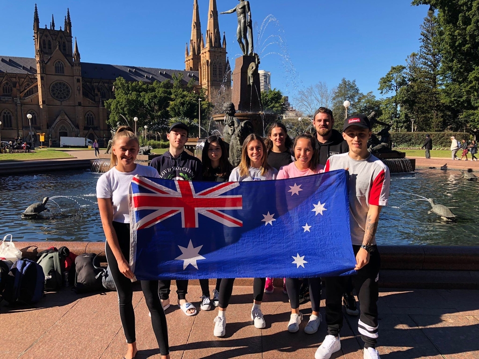 Groupe tenant un drapeau australien avec une fontaine et un bâtiment historique en arrière-plan.