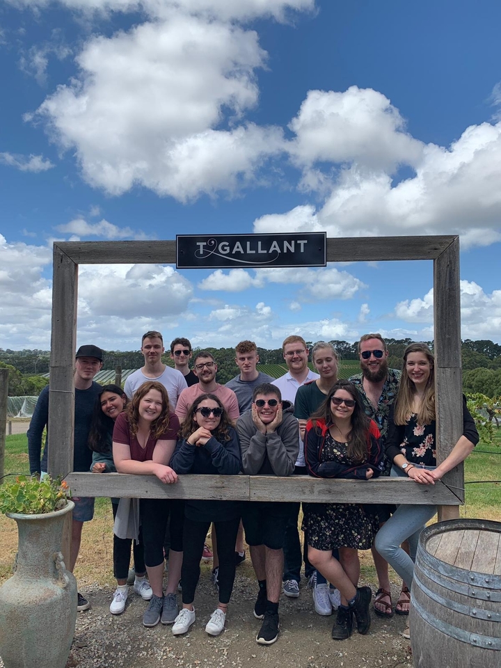 Group photo under a wooden sign with vineyards in the background.