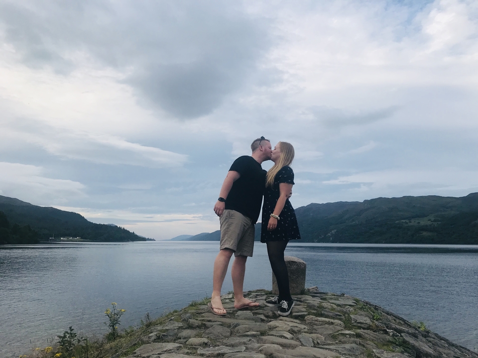 Couple kissing by a lake with mountains in the background.