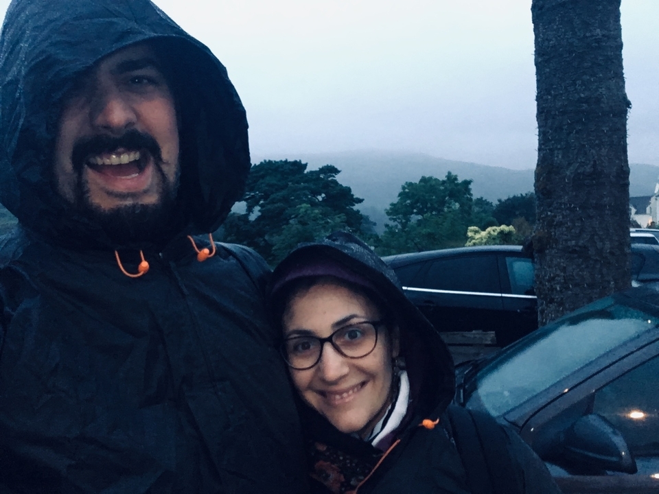 Two people bundled in raincoats smiling with a blurry, rainy background.
