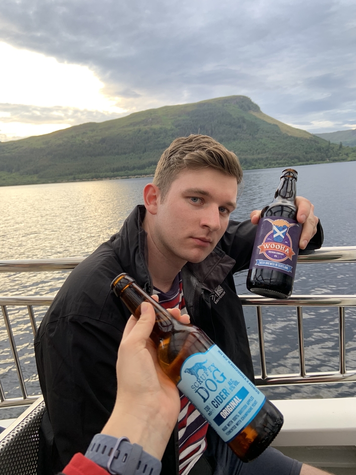 A person holding beer bottles on a boat with a scenic water backdrop.