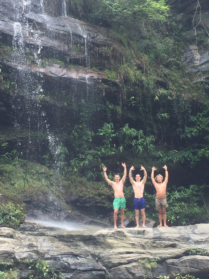 Trois personnes sous une cascade levant les bras, entourées d'une végétation luxuriante.