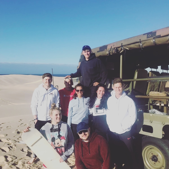 Group posing by a military vehicle on sand dunes.