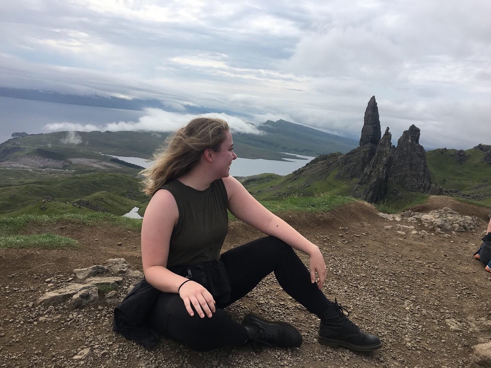 Person posing on a hillside with a view of rock formations and sea.