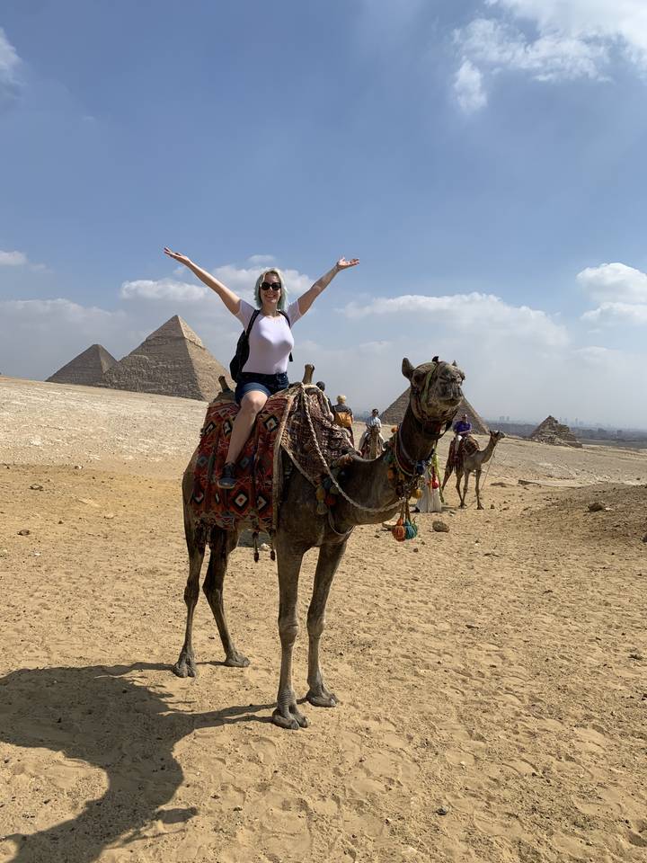 Person riding a camel with pyramids in the background.