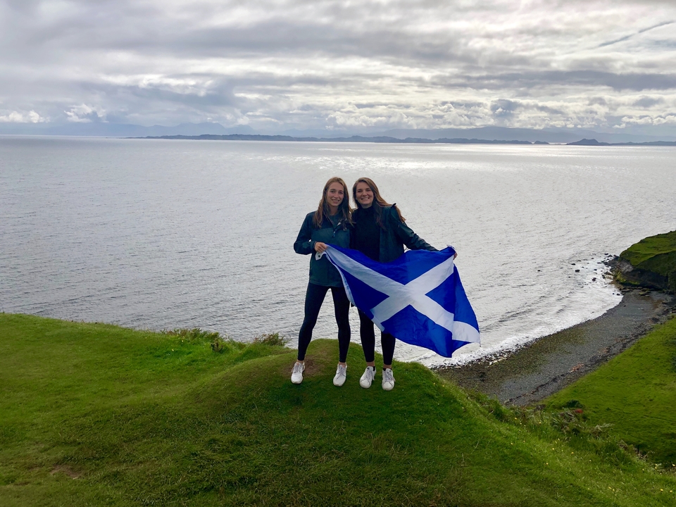 Two people holding a Scottish flag on a grassy hill overlooking the sea.