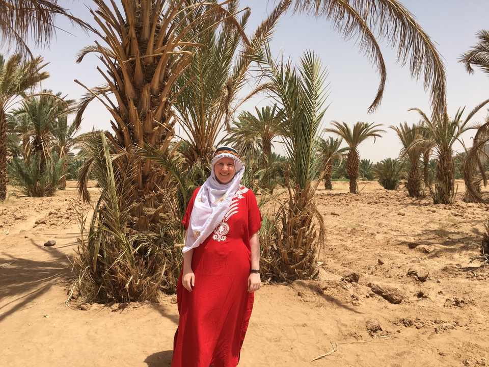 A woman wearing traditional attire standing among palm trees.