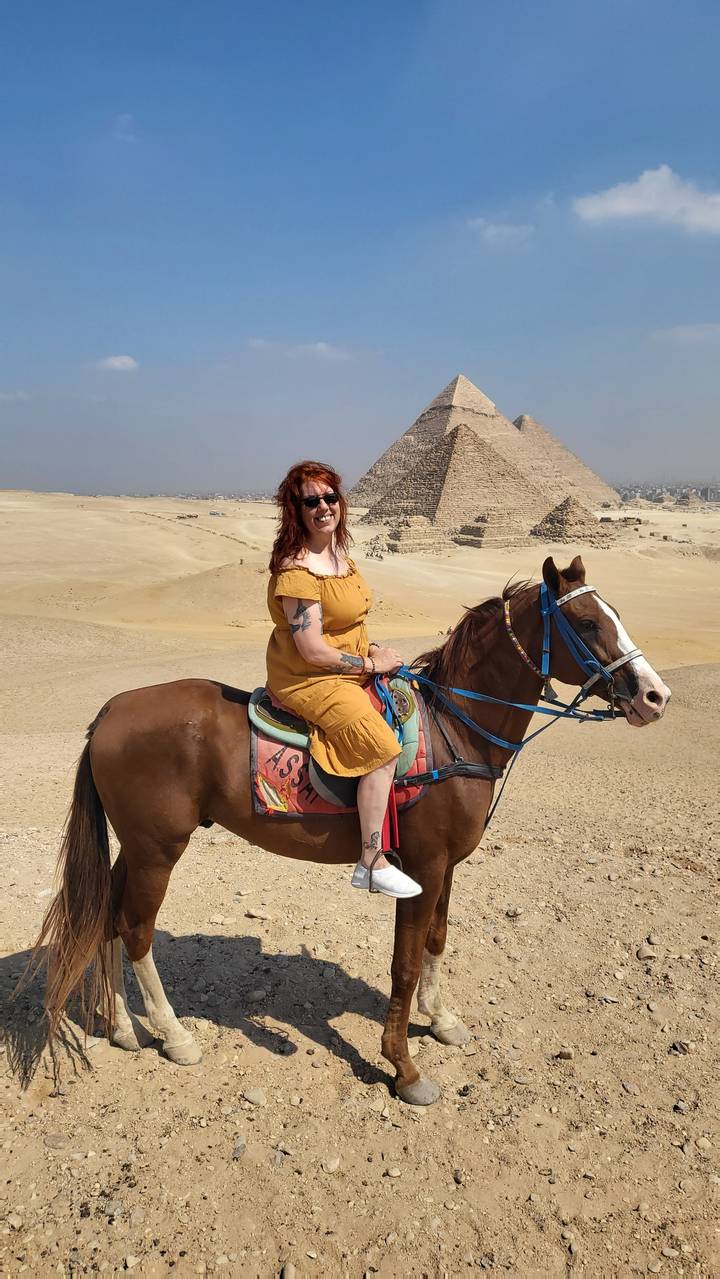 Person riding a horse in front of pyramids in the desert.