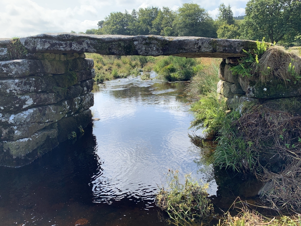 Stone clapper bridge over a river with surrounding greenery.