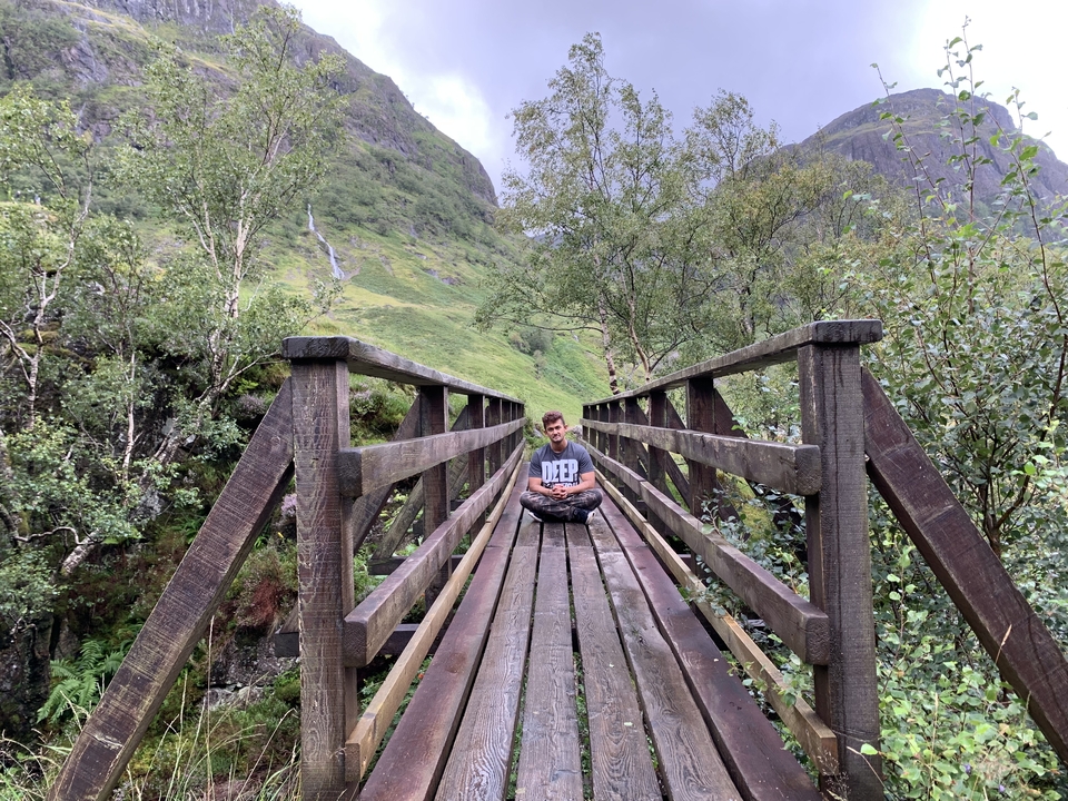 Person sitting on a wooden bridge in a lush setting.