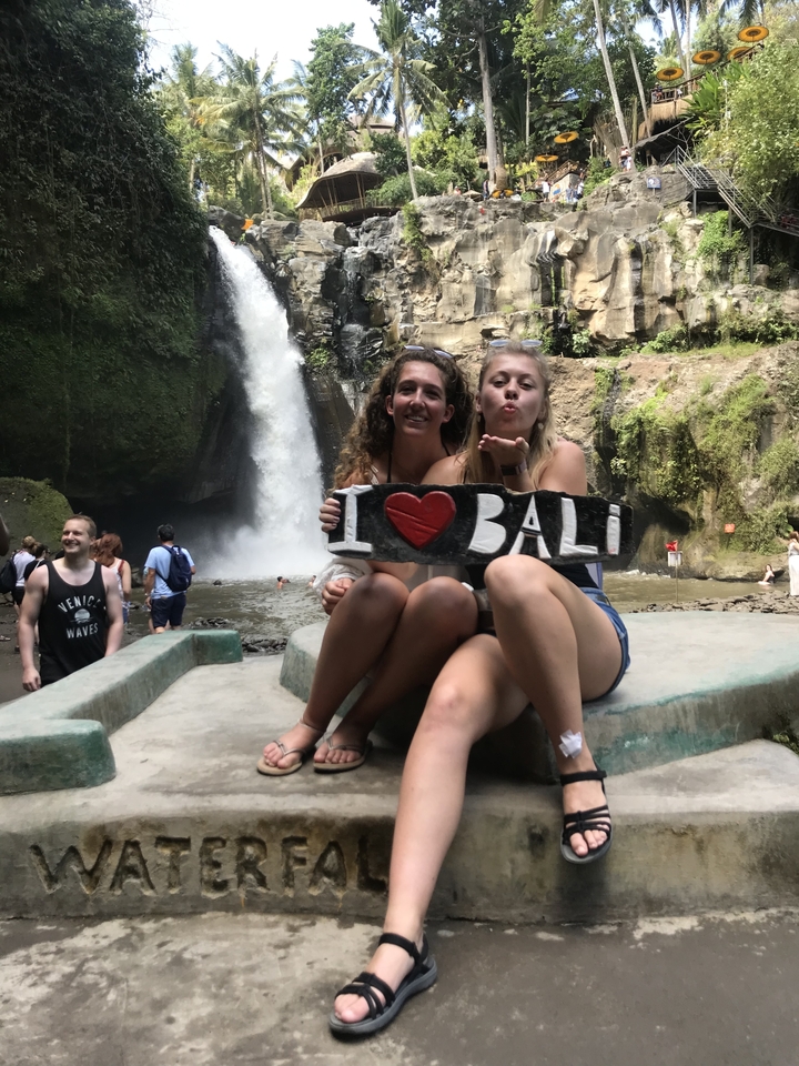 Two women holding an 'I Love Bali' sign in front of a waterfall.