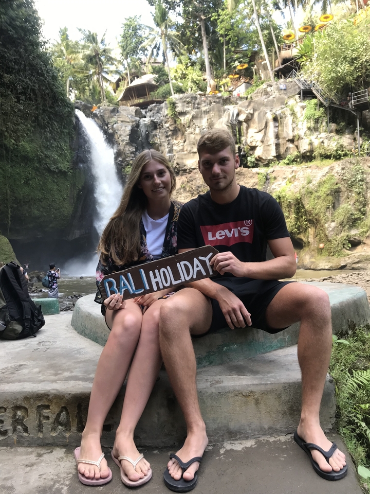 Couple holding 'Bali Holiday' sign in front of a waterfall.