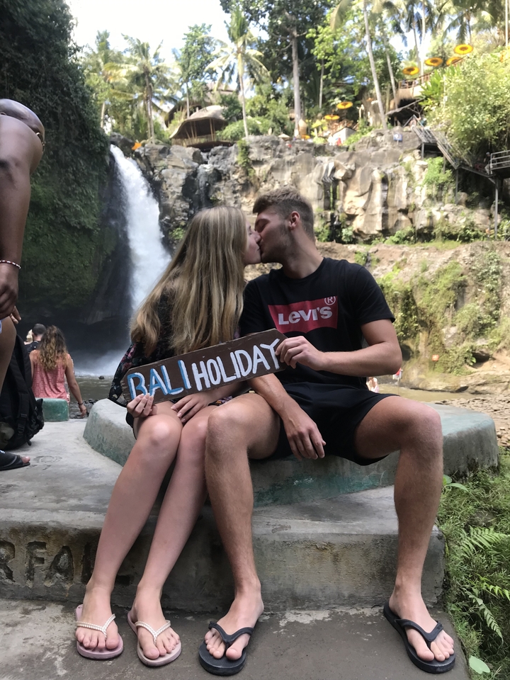 Couple kissing while holding 'Bali Holiday' sign in front of a waterfall.