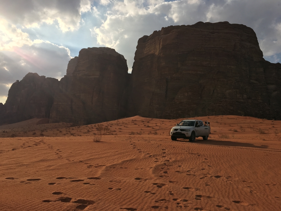 Une voiture garée sur un désert sablonneux avec de grandes formations rocheuses sous un ciel partiellement nuageux.
