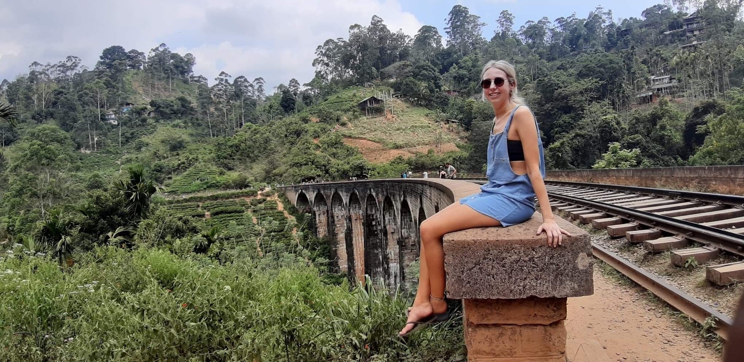 Woman sitting on a bridge with a lush landscape and train tracks.