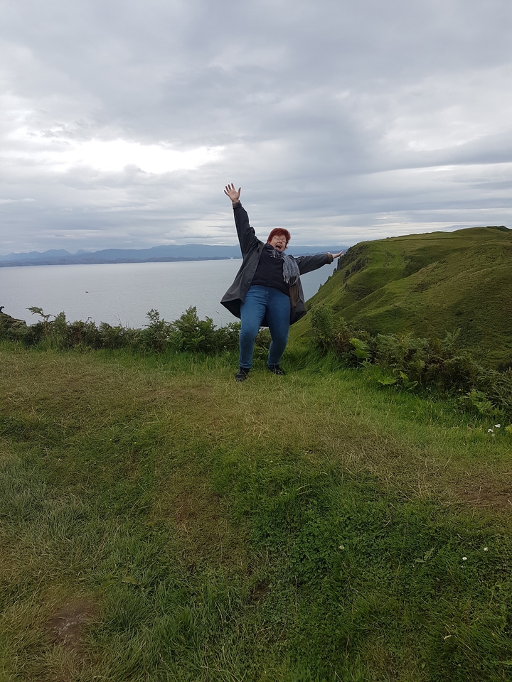 A person enthusiastically posing on a grassy hilltop overlooking the sea.