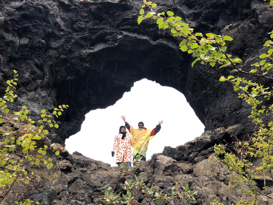 Two people standing under a large rock arch surrounded by green foliage.