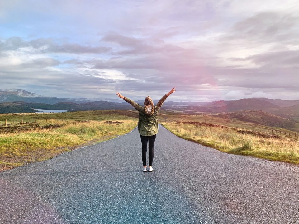Person standing on an open road with arms raised, surrounded by hills.