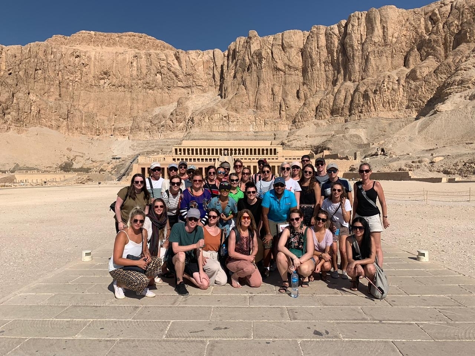 A group of tourists posing in front of the Temple of Hatshepsut with cliffs in the background.