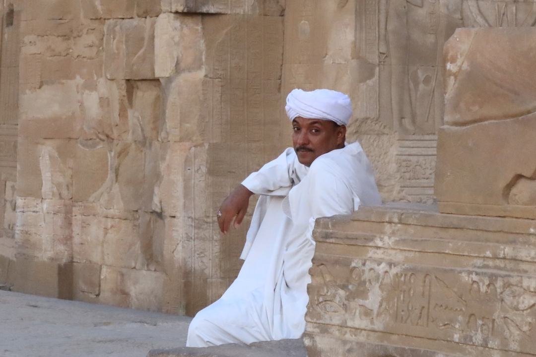 Homme en tenue traditionnelle assis contre un mur de pierre ancien.