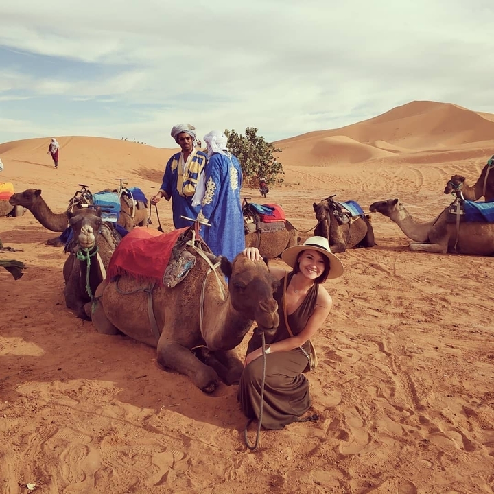 A person sitting next to a camel, with others in the background.