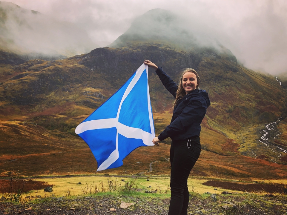 Person holding a Scottish flag in front of a mountainous landscape.