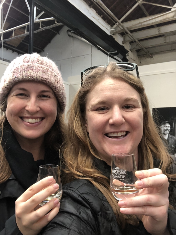 Deux femmes souriantes à l'intérieur, l'une tenant un verre.