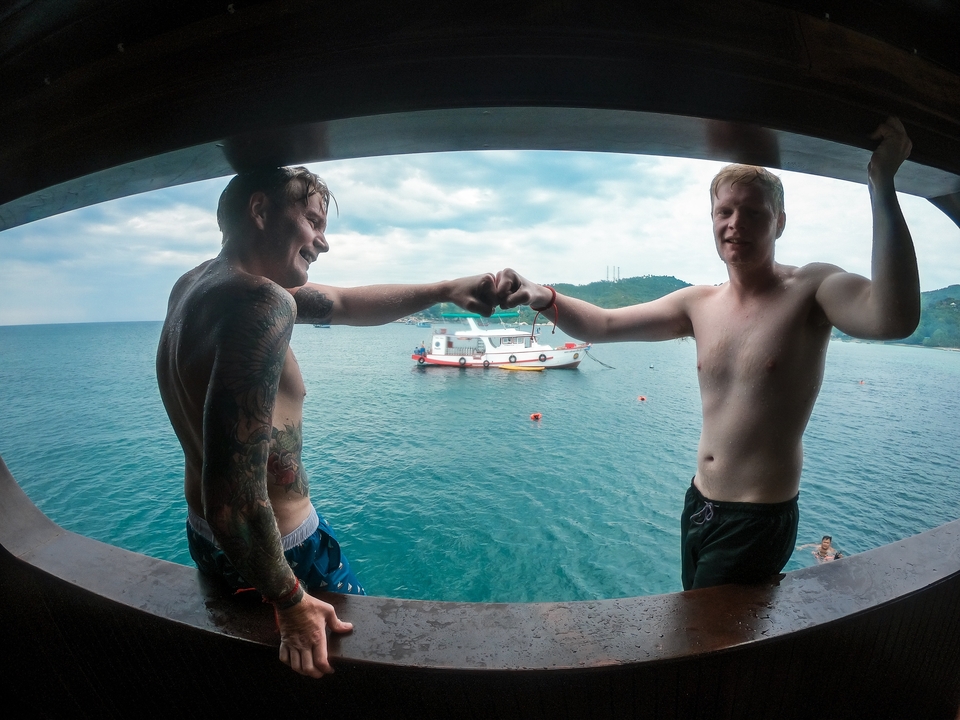 Two men fist-bumping on a boat with a scenic view of the ocean and a distant boat.