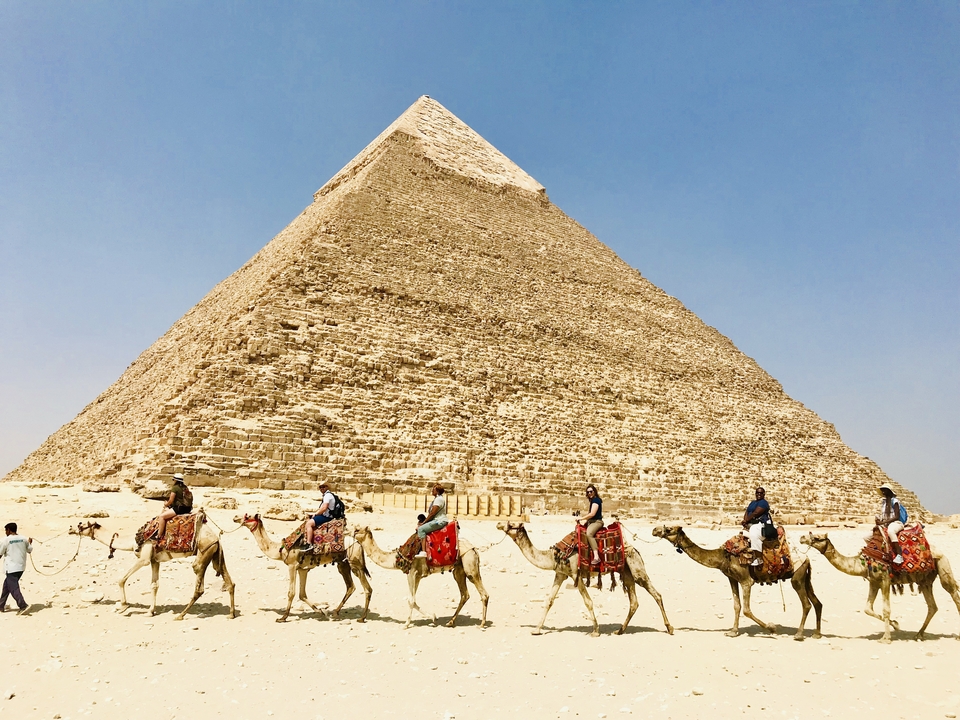 Camel riders passing by the Great Pyramid under a clear blue sky.