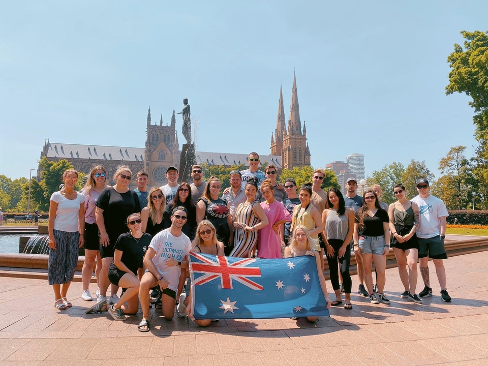 Groupe de personnes à l'extérieur tenant un drapeau australien, avec un bâtiment historique visible.