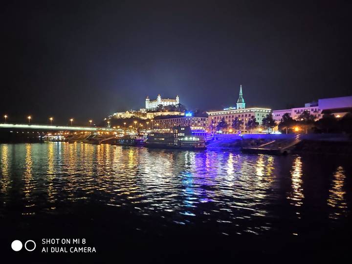 Paysage urbain nocturne avec des bâtiments illuminés et un pont se reflétant sur une rivière.