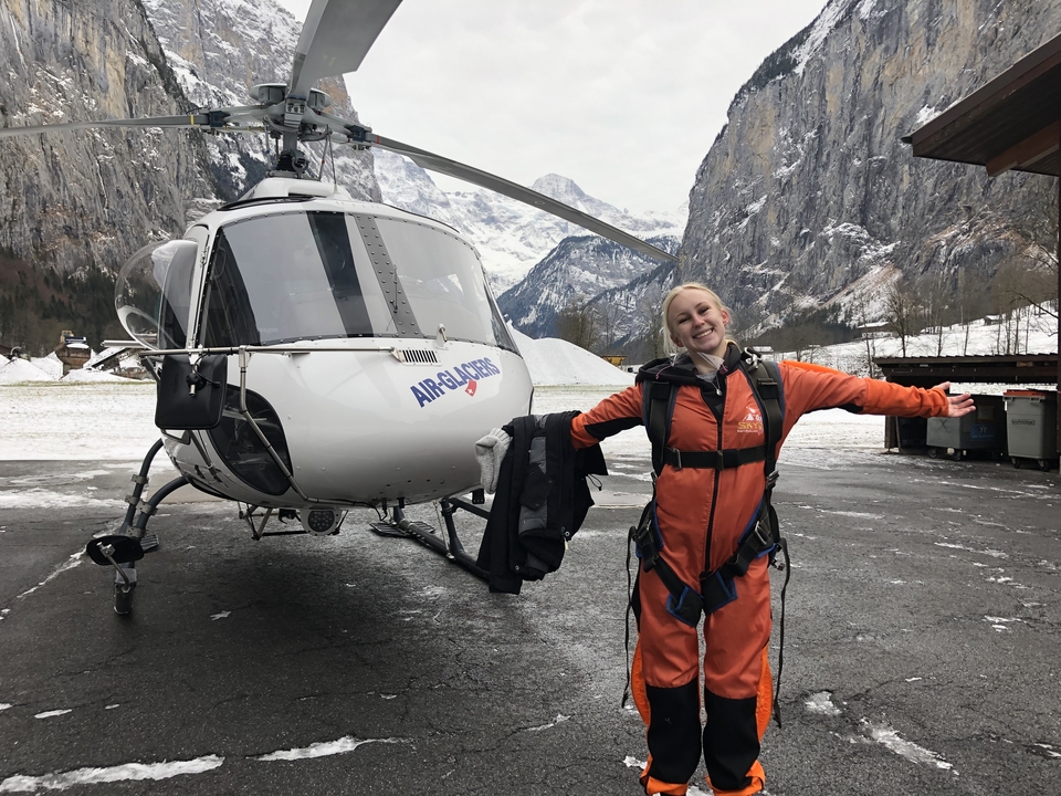 Person posing in front of a helicopter with mountainous background.