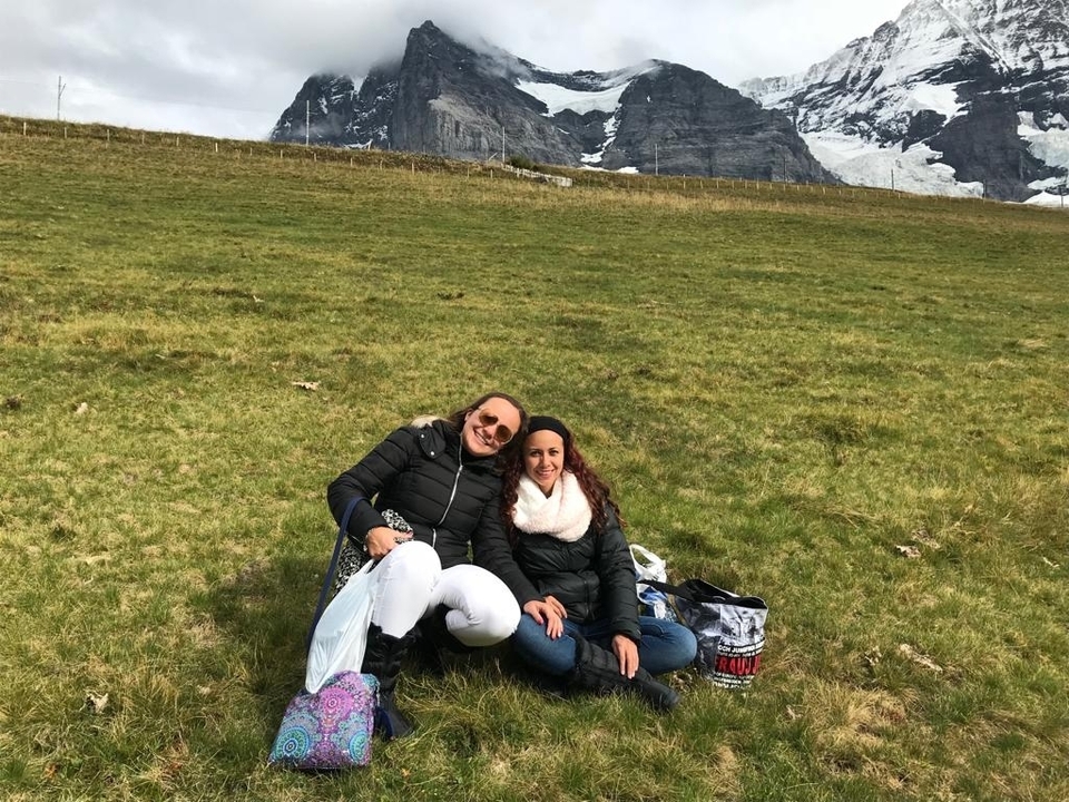 Two women sitting on grass in front of alpine mountains.