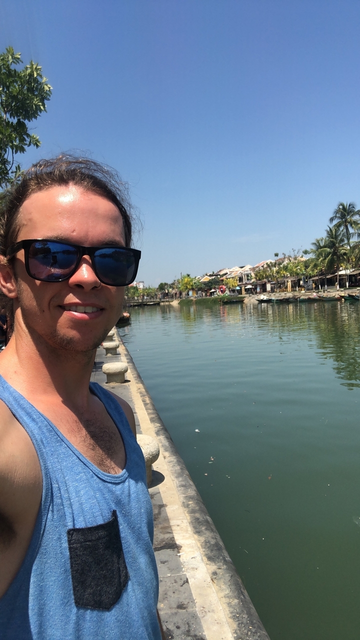 Person posing by a canal with boats and buildings in the background.