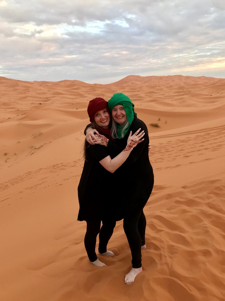 Two women dressed in traditional Moroccan attire standing in the desert.