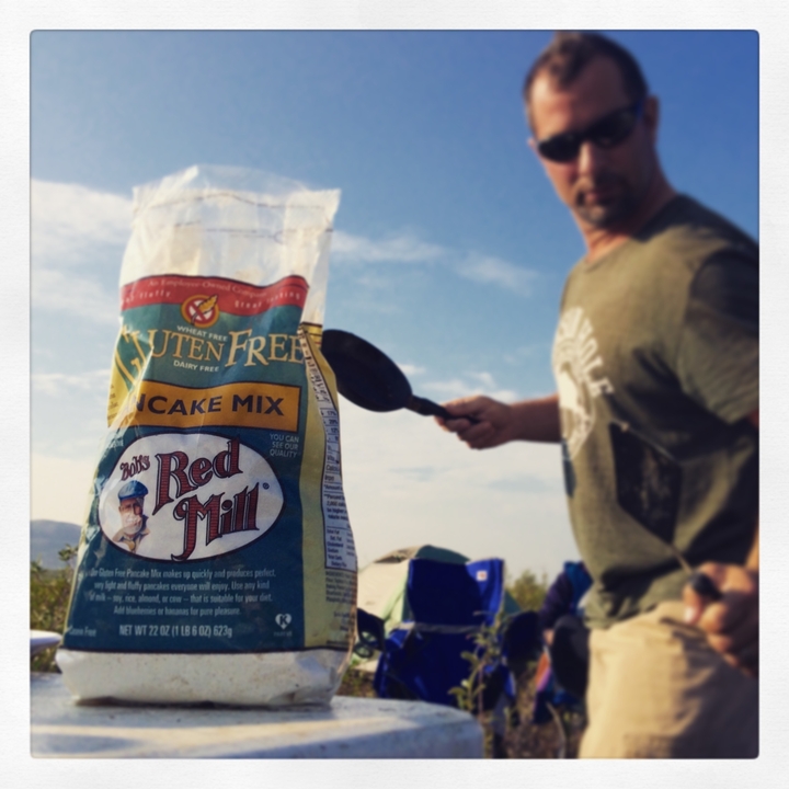 A person holding pancake mix beside a campfire.