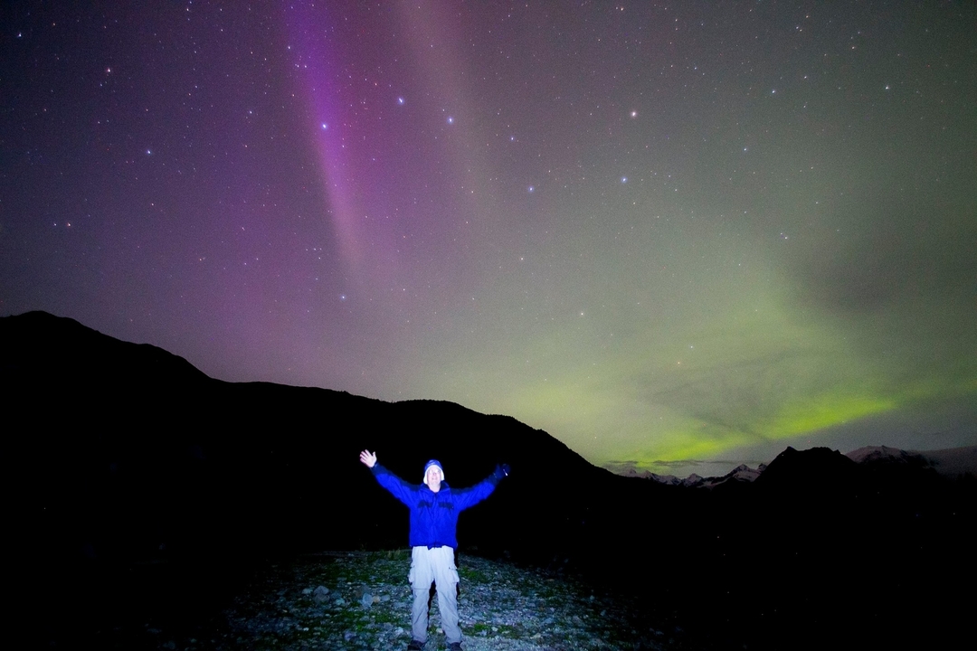 Person celebrating under a colorful aurora in the night sky.