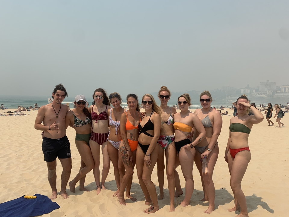 Group of friends in swimwear posing on a sandy Australian beach with hazy ocean backdrop.
