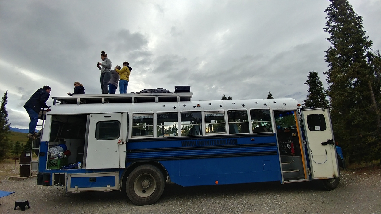 People on a large blue bus, some on the roof in a rural area.