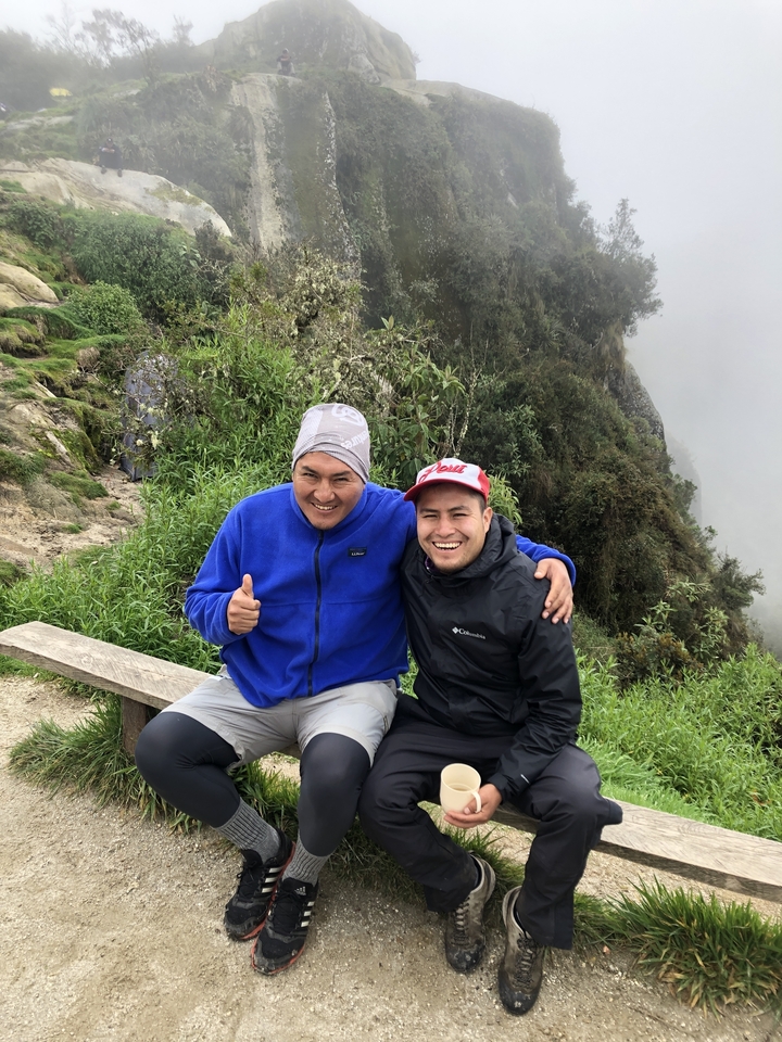Deux randonneurs souriants assis sur un banc dans un paysage luxuriant.