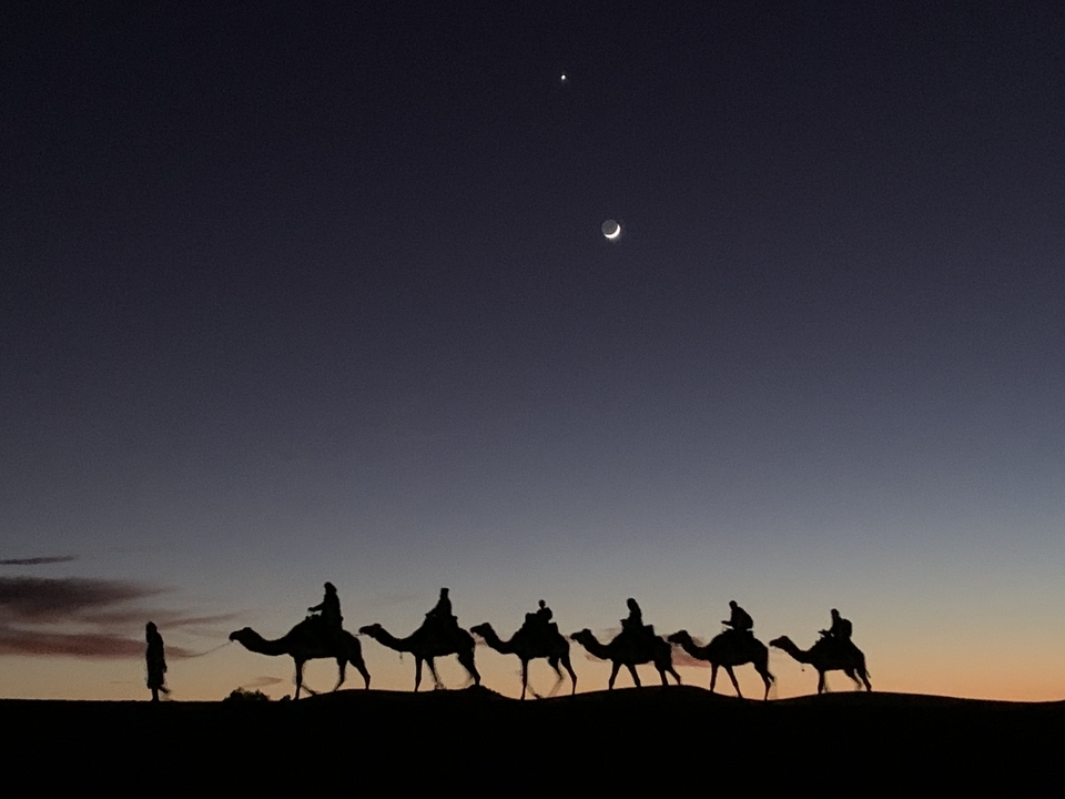 Silhouettes of camels and riders against a twilight sky with a crescent moon.