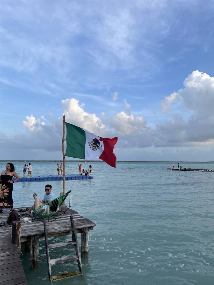 A Mexican flag on a pole near a beach with people and the ocean.