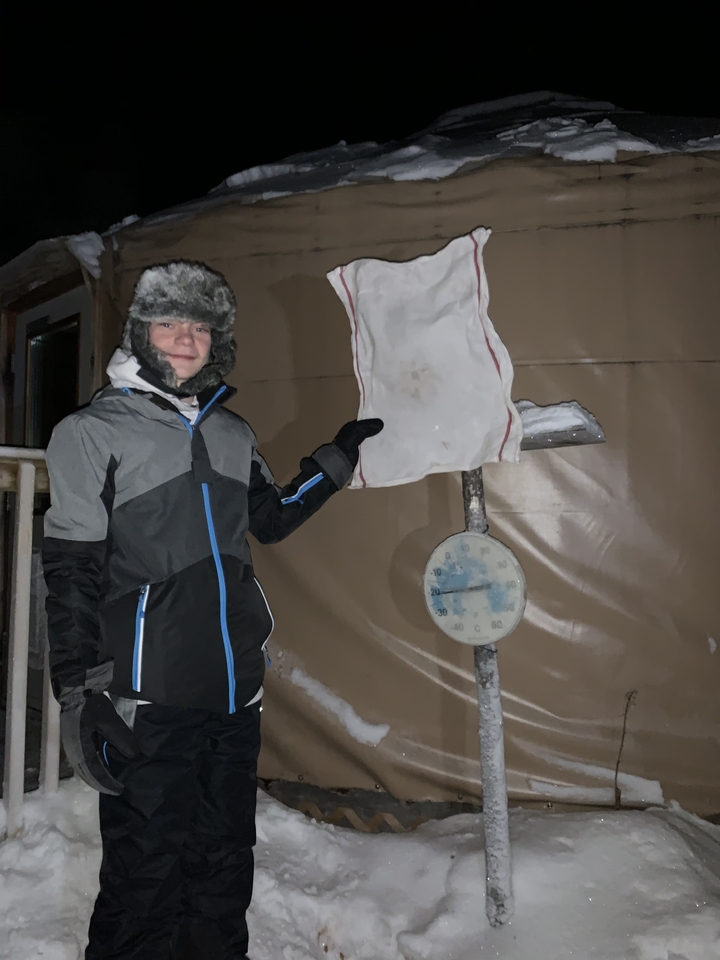 A person smiling and pointing to a snow-covered temperature gauge at night.