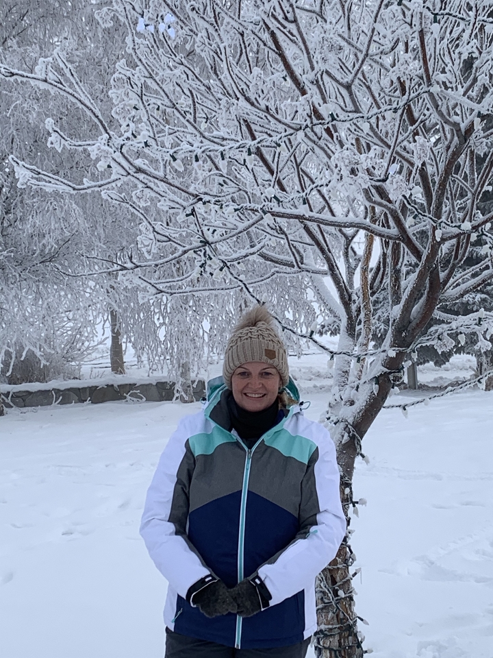 A person smiling under a snowy tree, wearing a winter jacket and beanie.