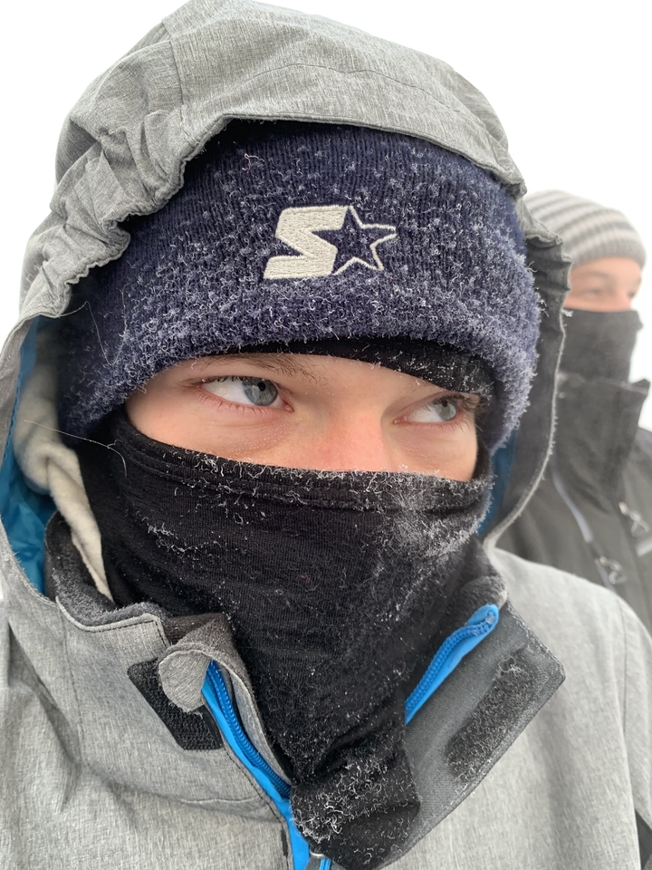 Close-up of a person's face bundled up for winter in front of a snowy background.