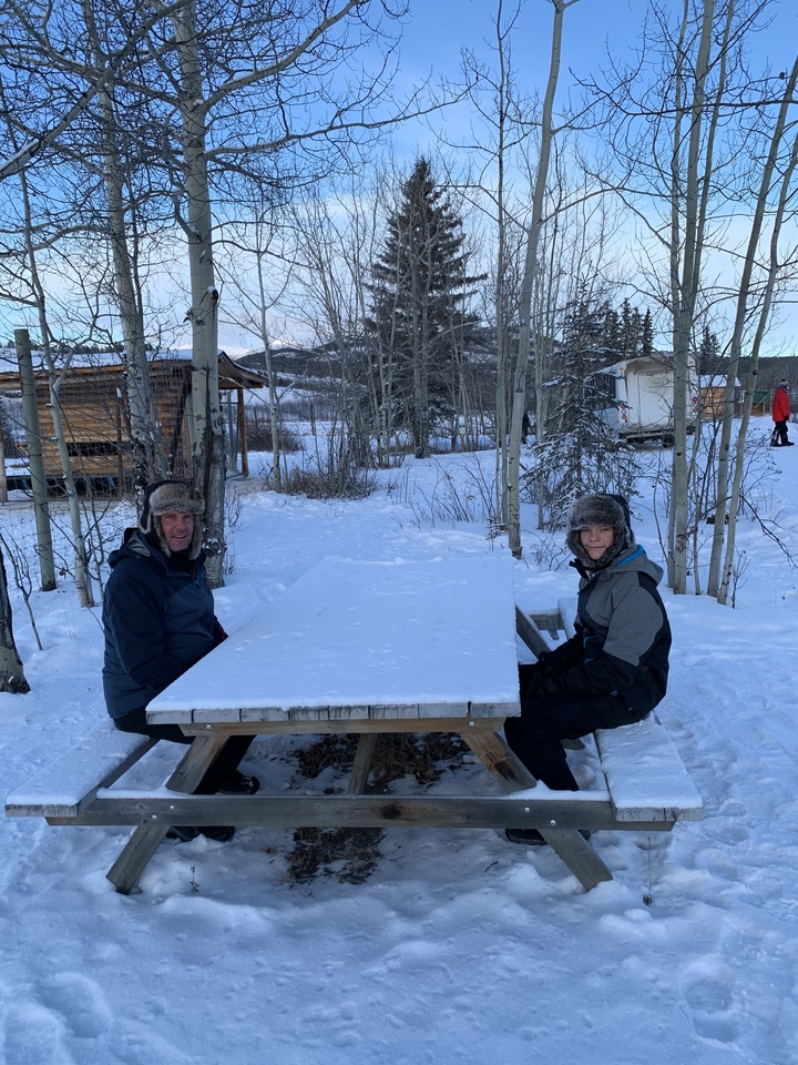 Two people sitting at a snow-covered picnic table in a wintry landscape.