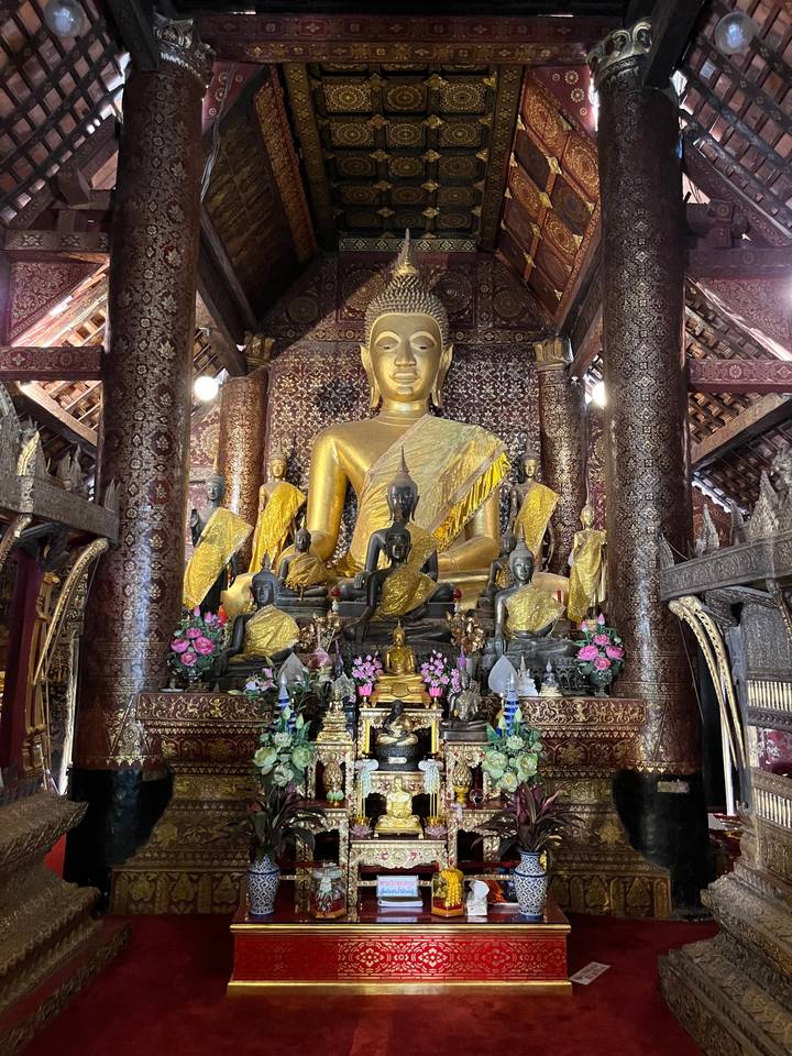 Statue de Bouddha dorée complexe entourée de statues plus petites et d'arrangements floraux.