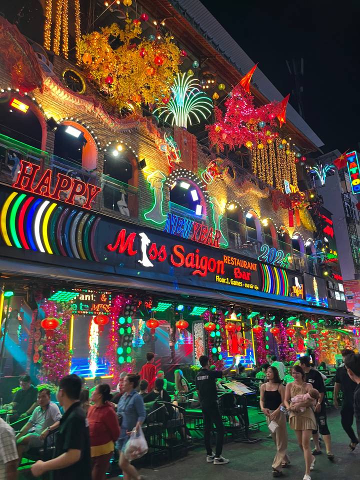 Scène nocturne animée devant un restaurant avec des enseignes au néon brillantes et des foules.
