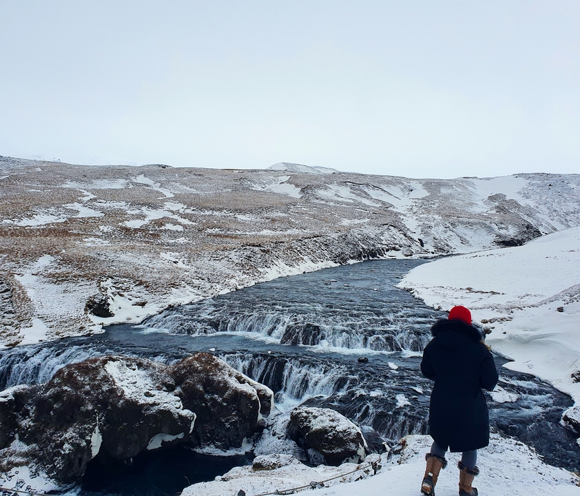 Person in winter attire standing by a snowy river with mountains.
