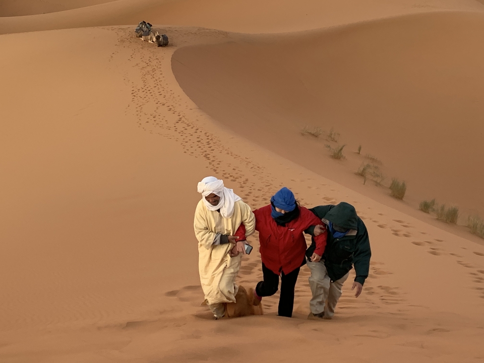 Three people walking up a sand dune in a desert.